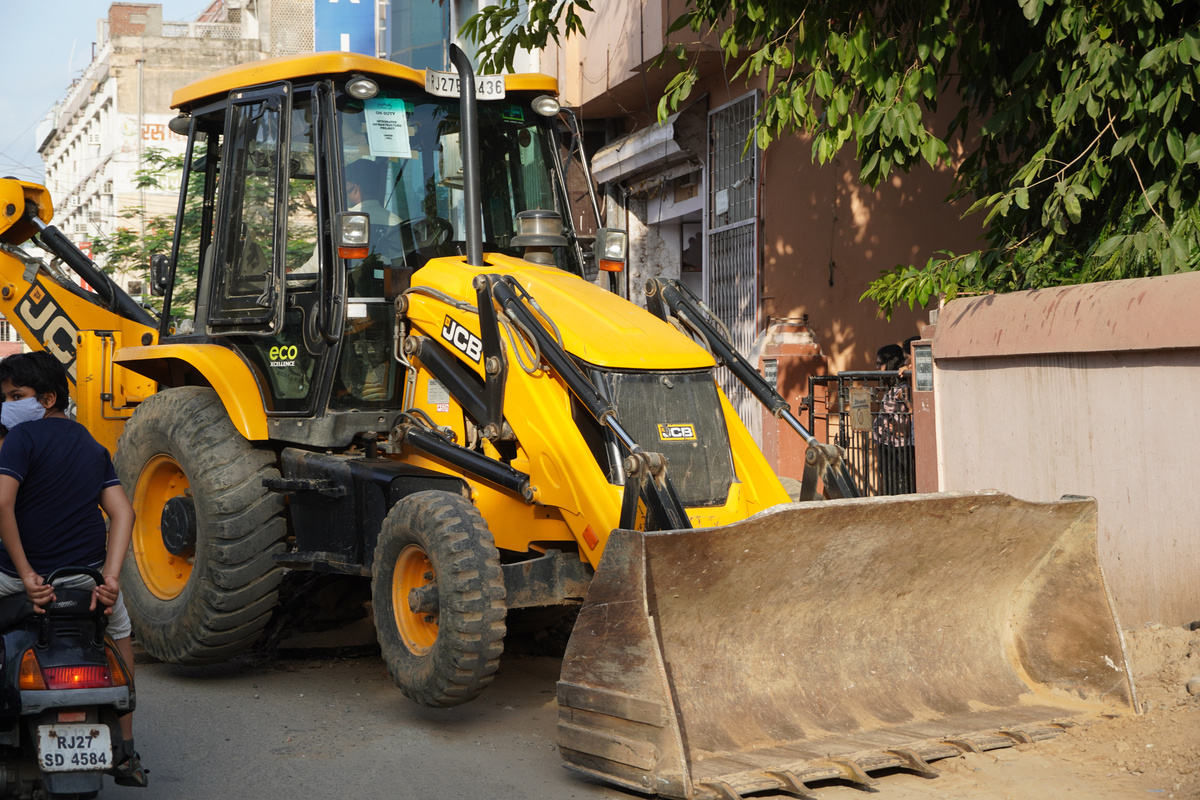 JCB earth moving machine working on a road. JCB bucket loader is repairing a section of a dirt road. Road is expanding in residential quarter. Theme of road construction. : Udaipur India - June 2020